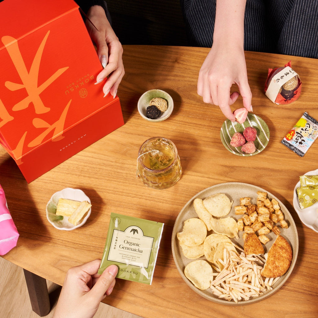 Three people sit around a wooden table enjoying Japanese snacks from the Bokksu Snack Box: Seasons of Japan, a red gift box, a glass of tea, and a packet labeled “Organic Genmaicha.”.